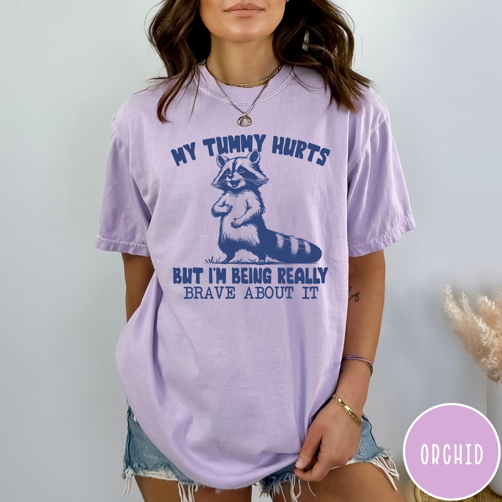 Comfort Colors tee in Orchid featuring graphic illustration of a standing raccoon with funny text about being brave