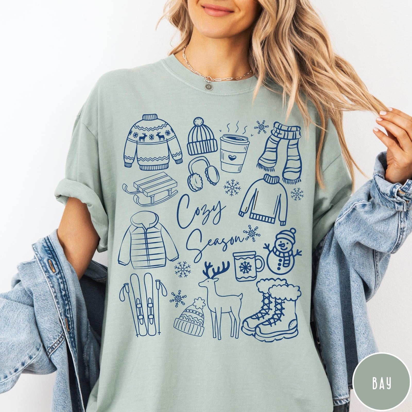 Comfort Colors tee in Bay featuring winter activities graphic with sweaters, sled, snowman, and seasonal items.