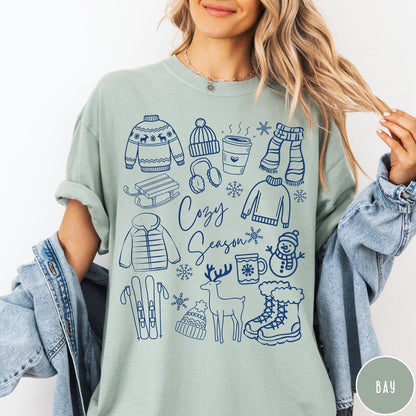 Comfort Colors tee in Bay featuring winter activities graphic with sweaters, sled, snowman, and seasonal items.