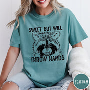Comfort Colors tee in Seafoam featuring graphic style raccoon illustration with text Sweet But Will Throw Hands