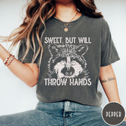 Comfort Colors tee in Pepper featuring graphic style raccoon illustration with text Sweet But Will Throw Hands