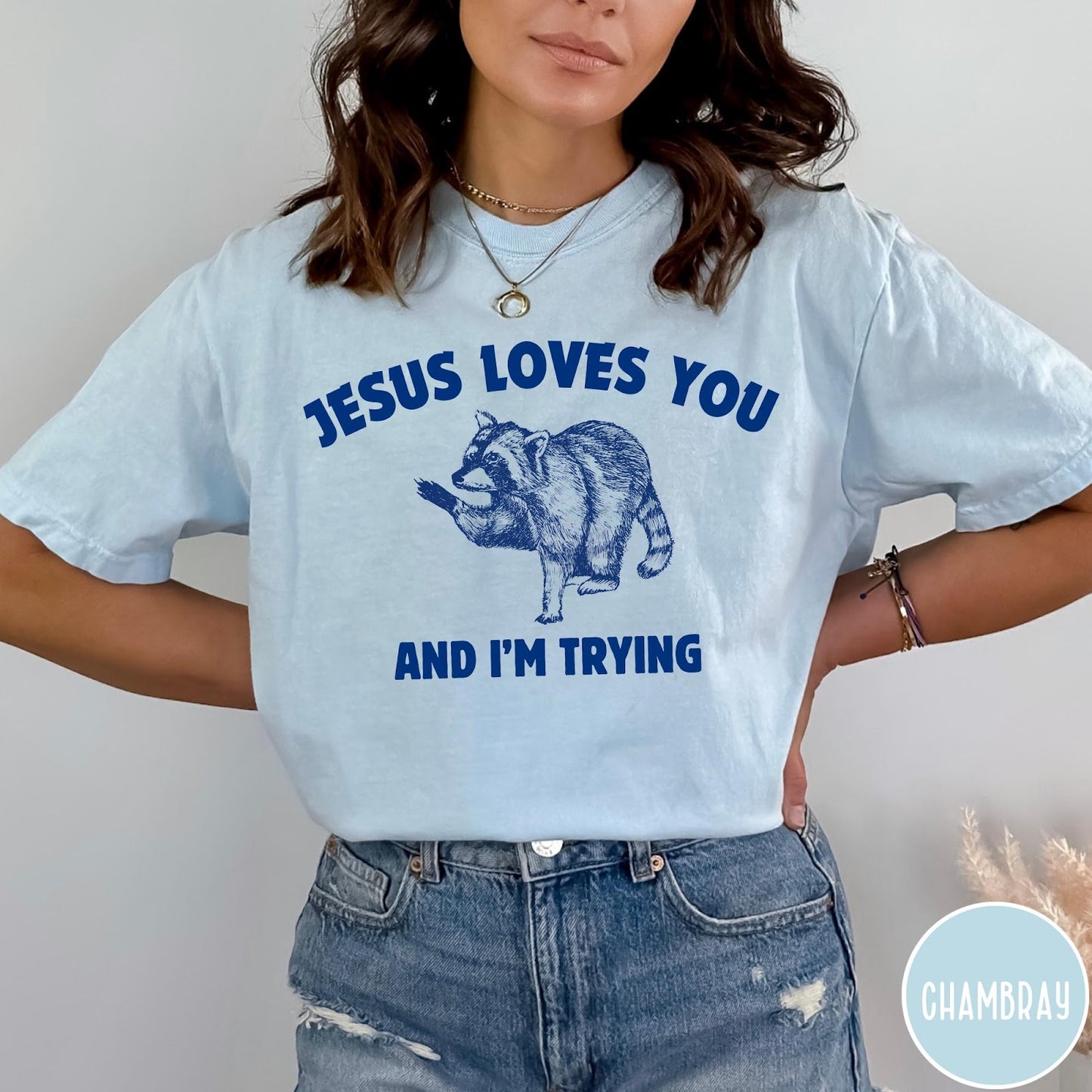 Comfort Colors tee in Chambray featuring graphic style raccoon illustration with text Jesus Loves You And I'm Trying