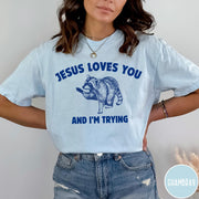 Comfort Colors tee in Chambray featuring graphic style raccoon illustration with text Jesus Loves You And I'm Trying