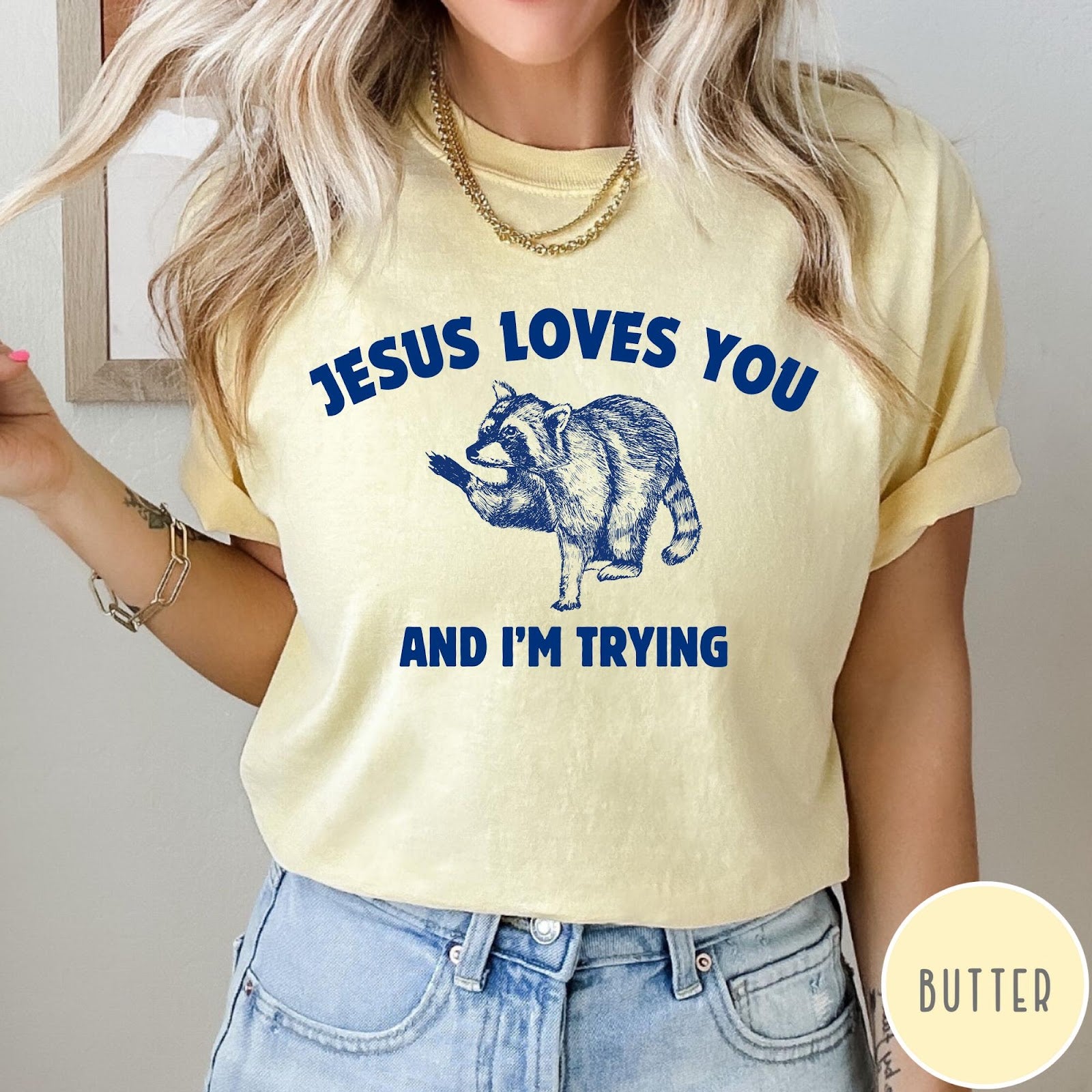 Comfort Colors tee in Butter featuring graphic style raccoon illustration with text Jesus Loves You And I'm Trying