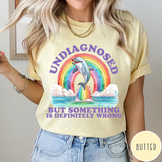 Comfort Colors tee in Butter featuring graphic style illustration of a dolphin jumping through a colorful rainbow