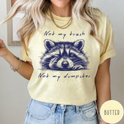 Comfort Colors tee in Butter featuring illustrated style graphic of a winking raccoon with text not my trash