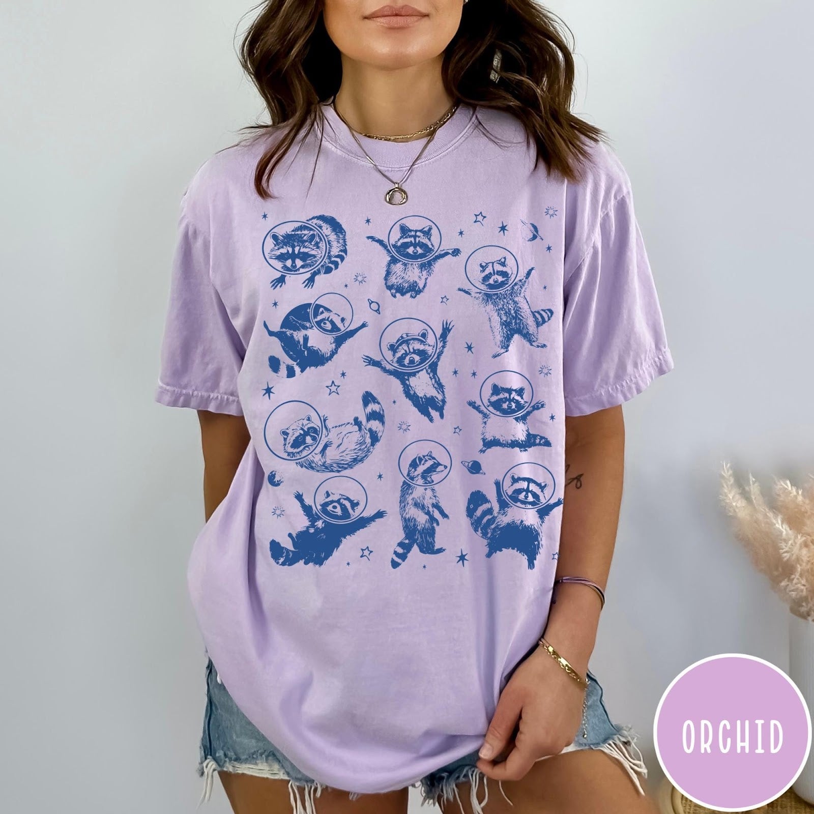 Comfort Colors tee in Orchid featuring whimsical space design with multiple raccoons wearing glass astronaut helmets and stars