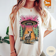 Comfort Colors tee in Ivory featuring graphic style cow being abducted by a UFO in the forest