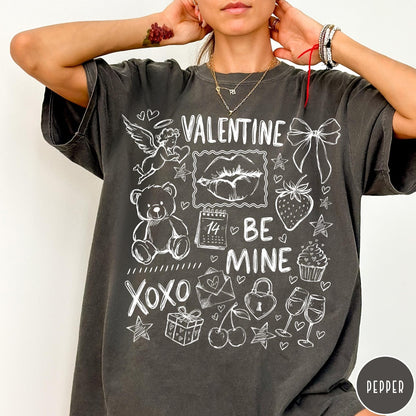 Comfort Colors tee in Pepper featuring sketch style Valentine's Day collage graphic with cupid, bear, and hearts