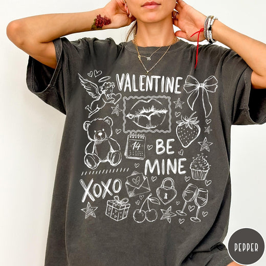 Comfort Colors tee in Pepper featuring sketch style Valentine's Day collage graphic with cupid, bear, and hearts