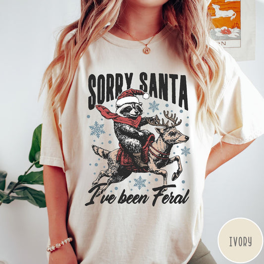 Comfort Colors tee in Ivory featuring festive graphic of a raccoon wearing a Santa hat riding a reindeer