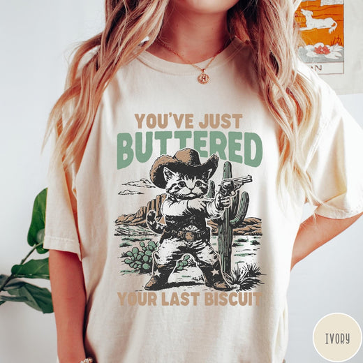Comfort Colors tee in Ivory featuring western graphic design of a cat wearing a cowboy hat and gun.