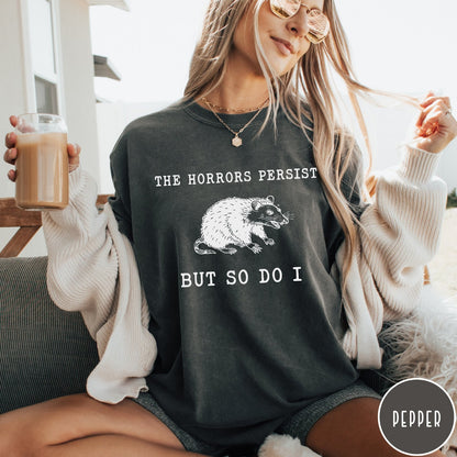 Comfort Colors tee in Pepper featuring illustrative style opossum graphic with text The Horrors Persist But So Do I