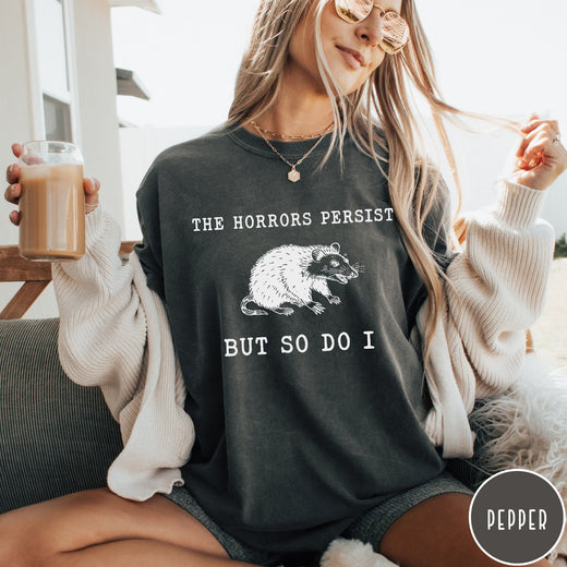 Comfort Colors tee in Pepper featuring illustrative style opossum graphic with text The Horrors Persist But So Do I