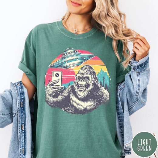 Comfort Colors tee in Light Green featuring retro graphic design of Bigfoot taking a selfie with a UFO