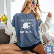 Comfort Colors tee in Blue Jean featuring illustrative style opossum graphic with text The Horrors Persist But So Do I