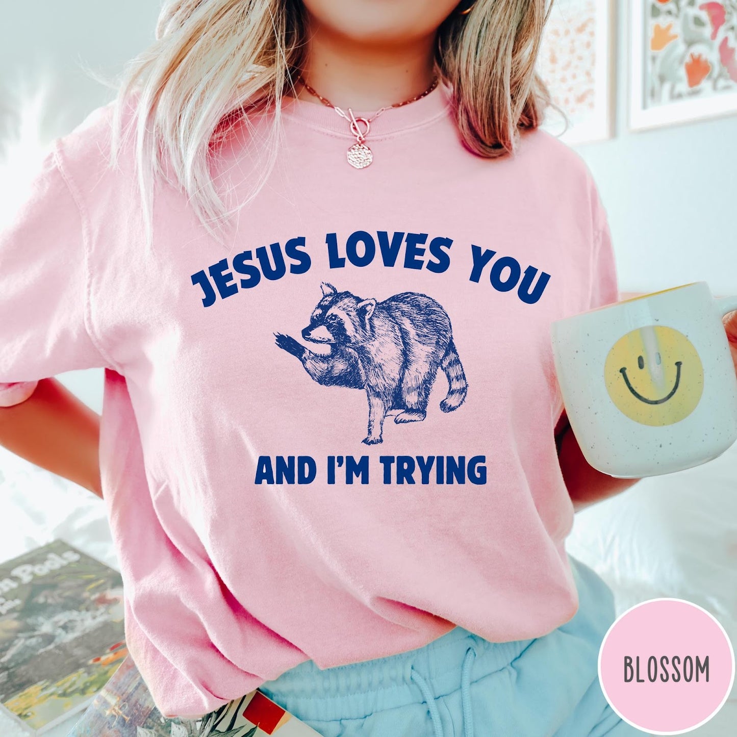 Comfort Colors tee in Blossom featuring graphic style raccoon illustration with text Jesus Loves You And I'm Trying
