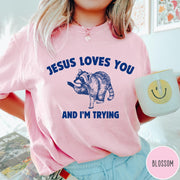 Comfort Colors tee in Blossom featuring graphic style raccoon illustration with text Jesus Loves You And I'm Trying