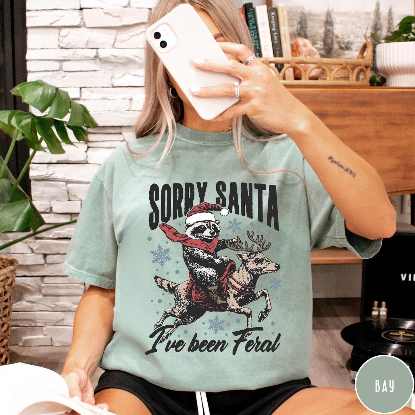 Comfort Colors tee in Bay featuring festive graphic of a raccoon wearing a Santa hat riding a reindeer