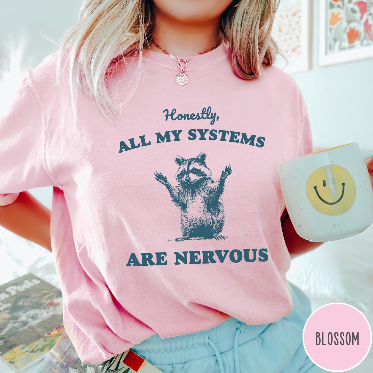 Comfort Colors tee in Blossom featuring graphic illustration of a nervous raccoon with arms raised in air