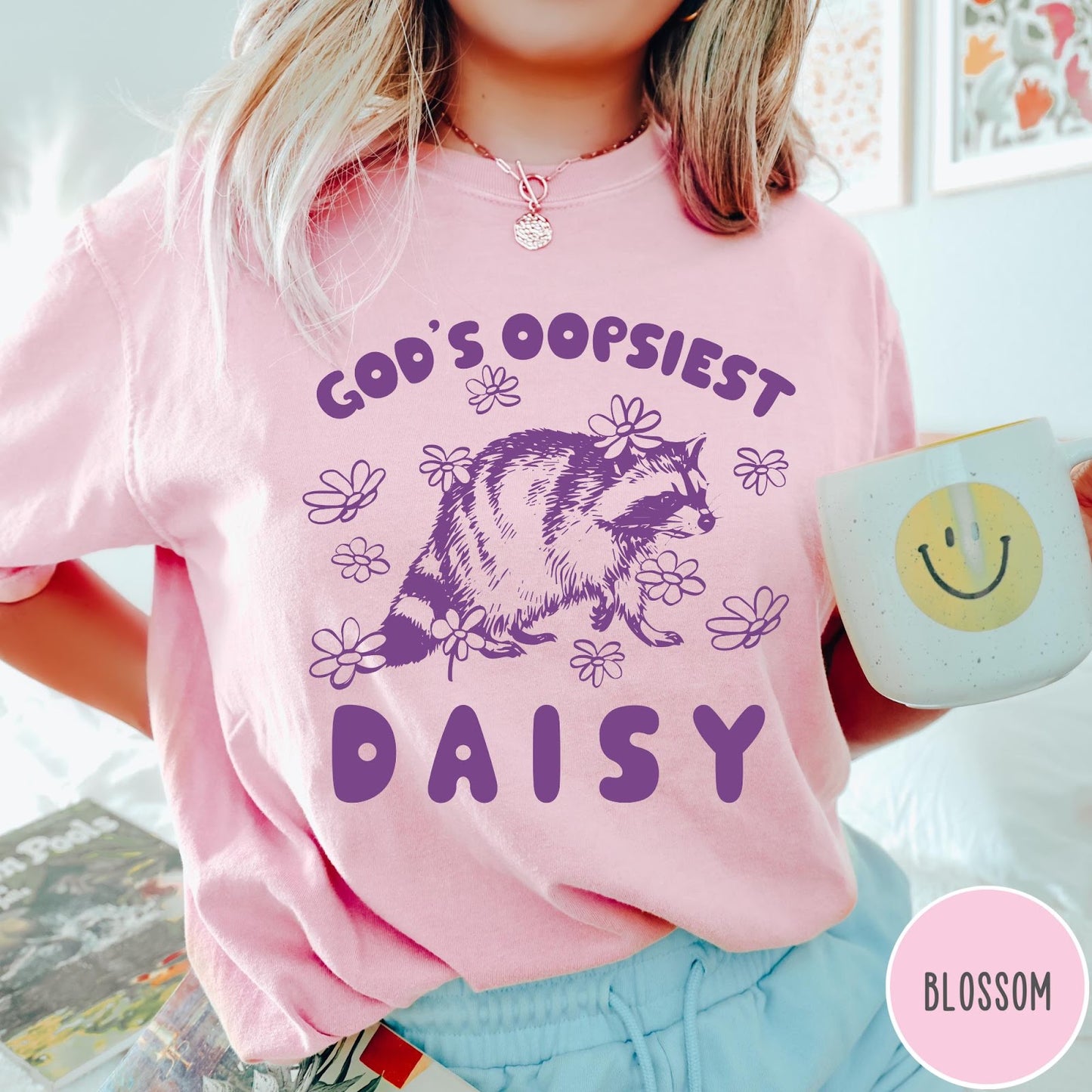 Comfort Colors tee in Blossom featuring whimsical raccoon graphic with daisy flowers and God's Oopsiest Daisy text