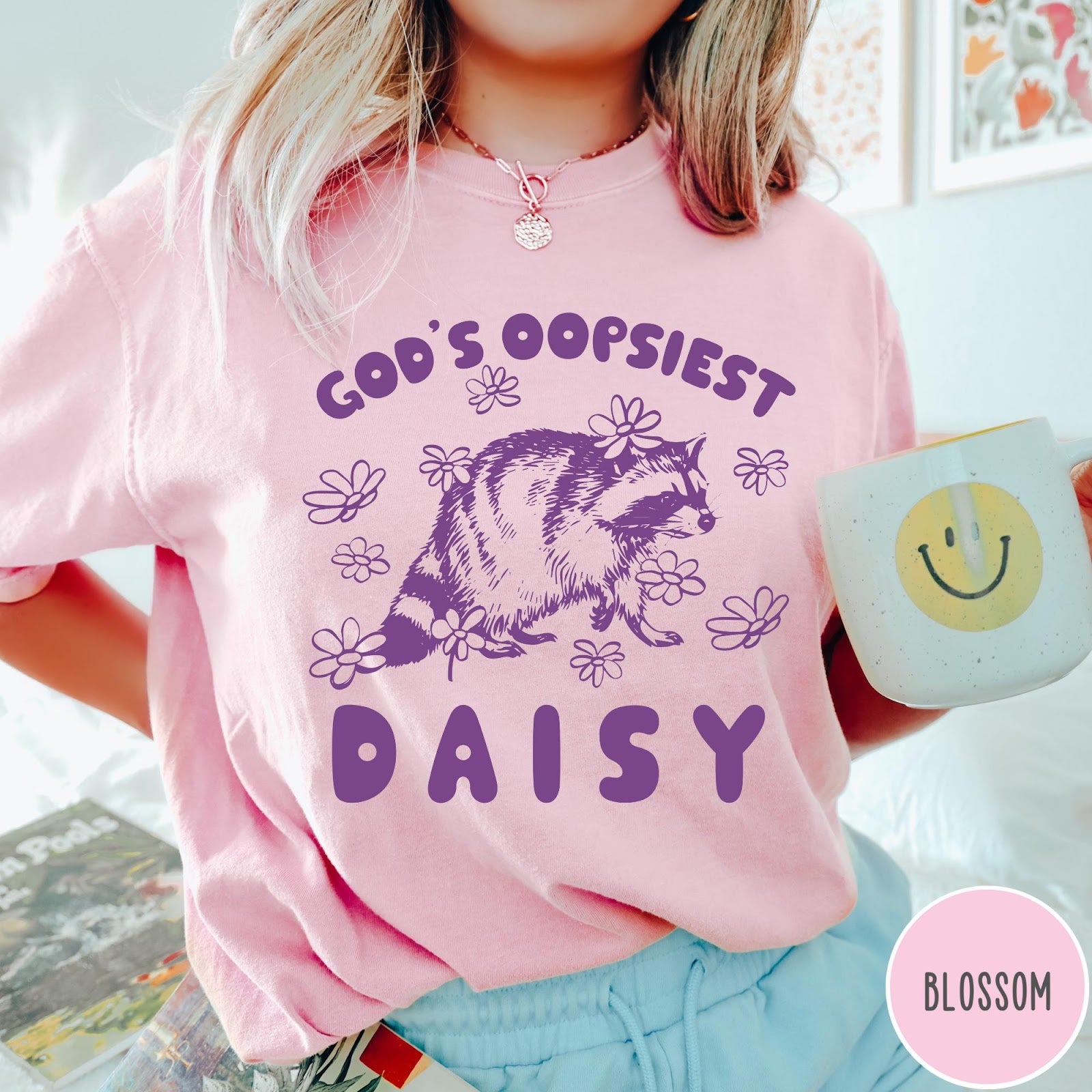 Comfort Colors tee in Blossom featuring whimsical raccoon graphic with daisy flowers and God's Oopsiest Daisy text