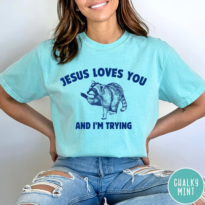Comfort Colors tee in Chalky Mint featuring graphic style raccoon illustration with text Jesus Loves You And I'm Trying