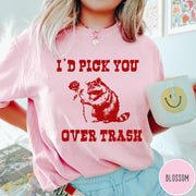 Comfort Colors tee in Blossom featuring a whimsical graphic of a raccoon holding a red rose.