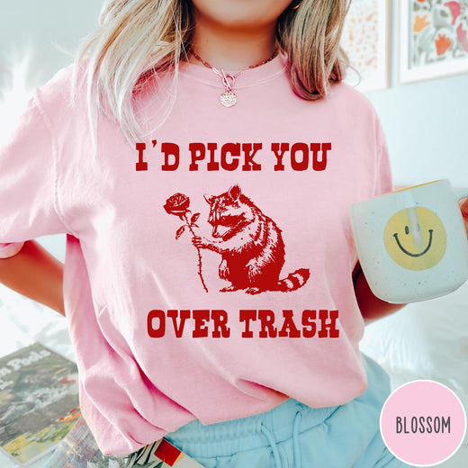 Comfort Colors tee in Blossom featuring a whimsical graphic of a raccoon holding a red rose.