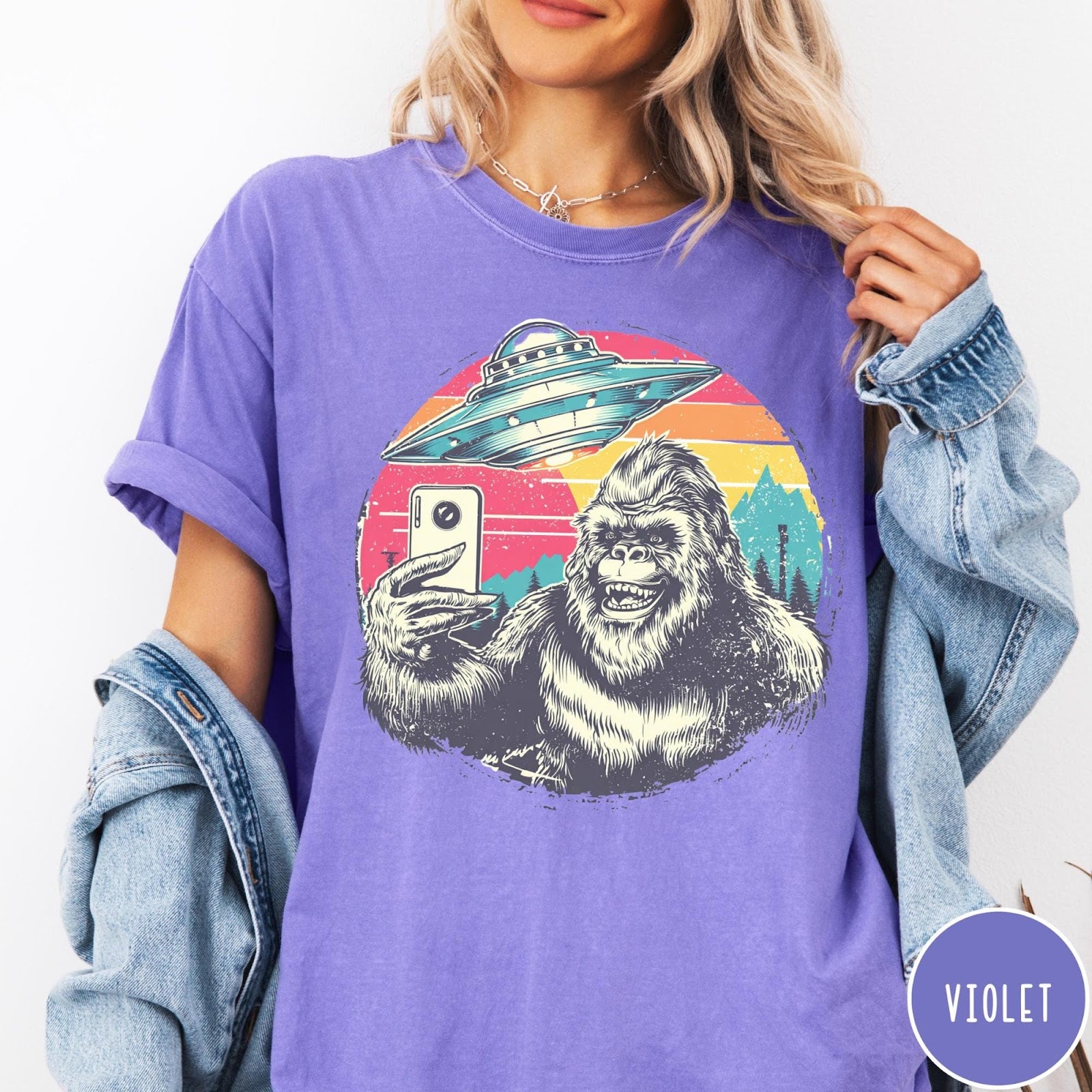 Comfort Colors tee in Violet featuring retro Bigfoot selfie graphic with a UFO and sunset mountain background
