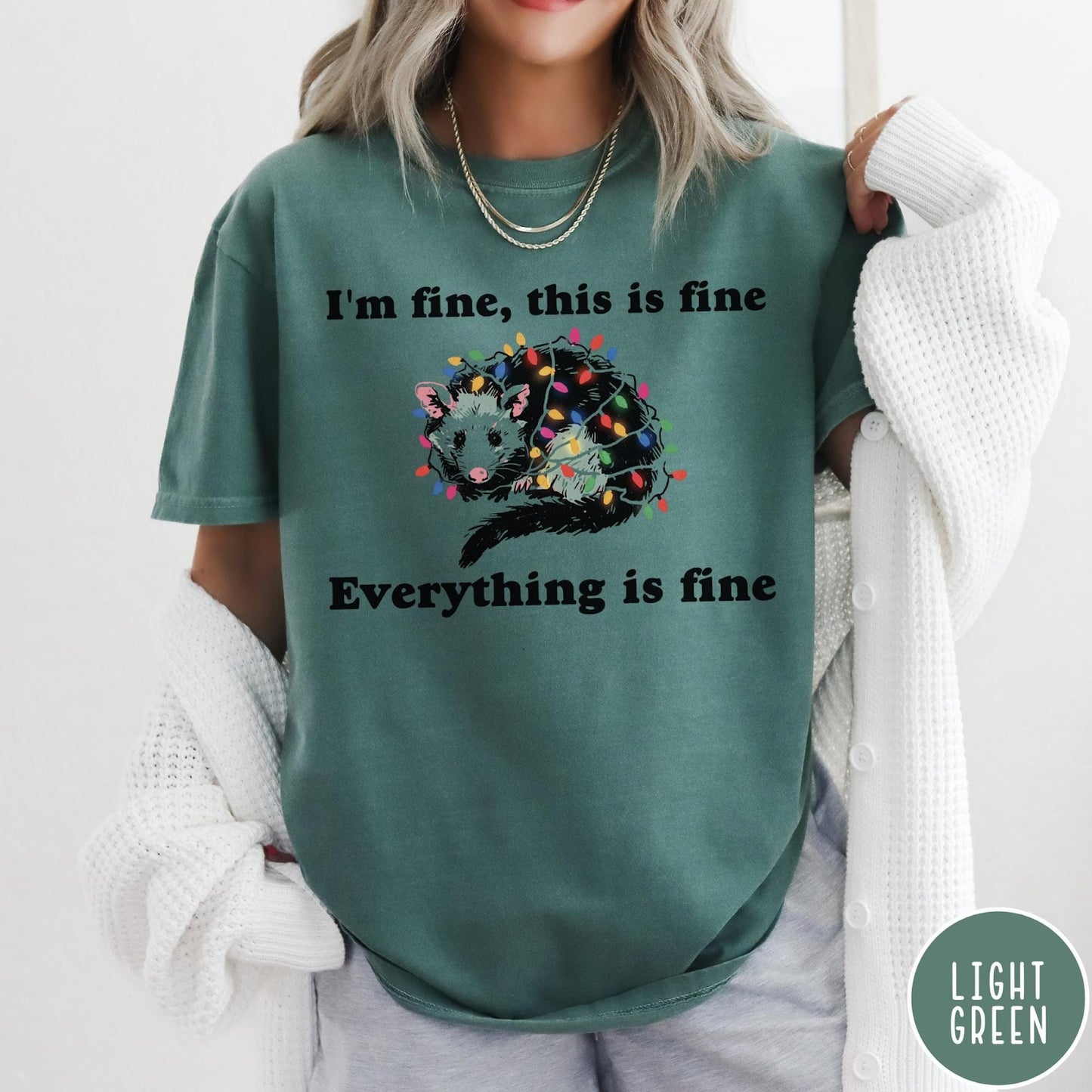 Comfort Colors tee in Light Green featuring graphic illustration of an opossum tangled in colorful Christmas lights