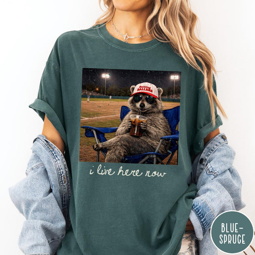 Comfort Colors tee in Blue Spruce featuring graphic style raccoon wearing sunglasses and baseball cap at a stadium
