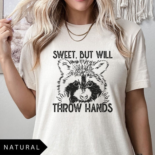 Comfort Colors tee in Natural featuring graphic style raccoon illustration with text Sweet, but will throw hands