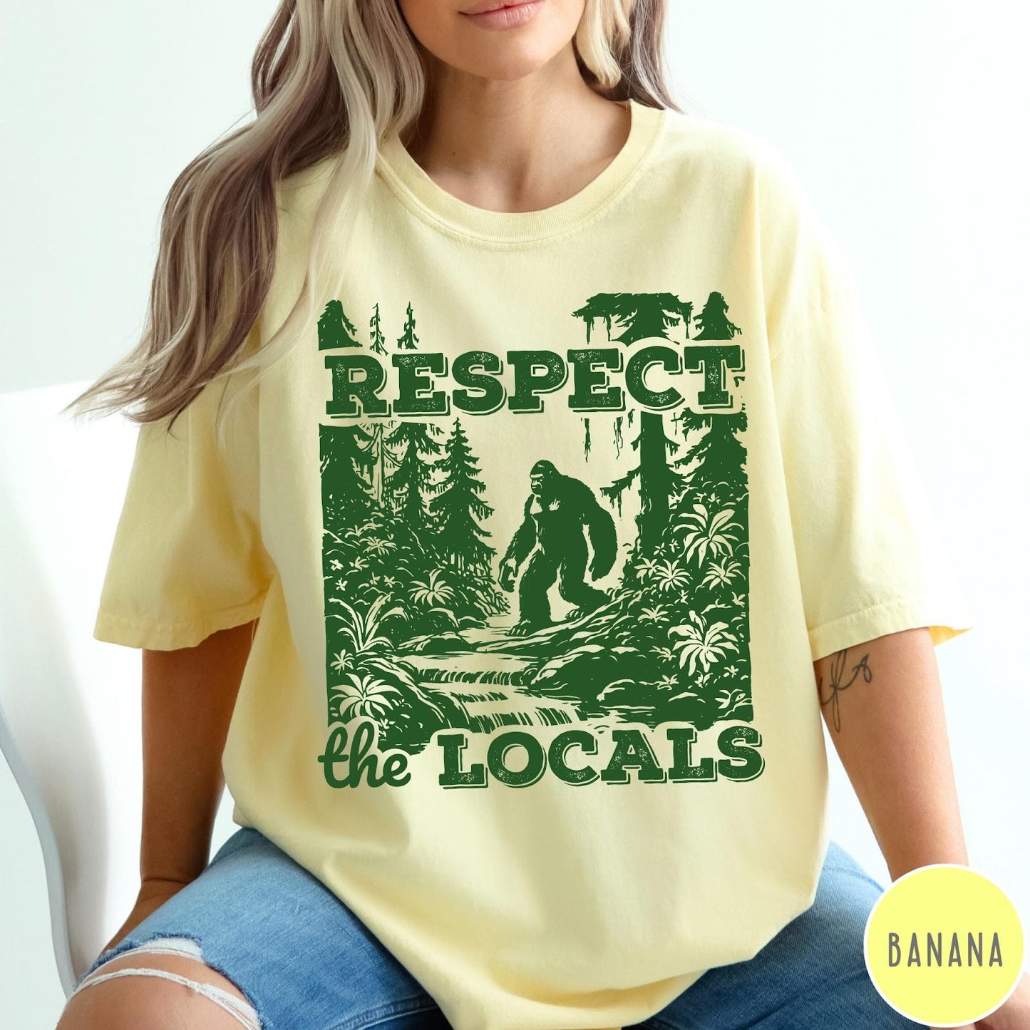 Comfort Colors tee in Banana featuring vintage style graphic of Bigfoot walking through a forest by a river