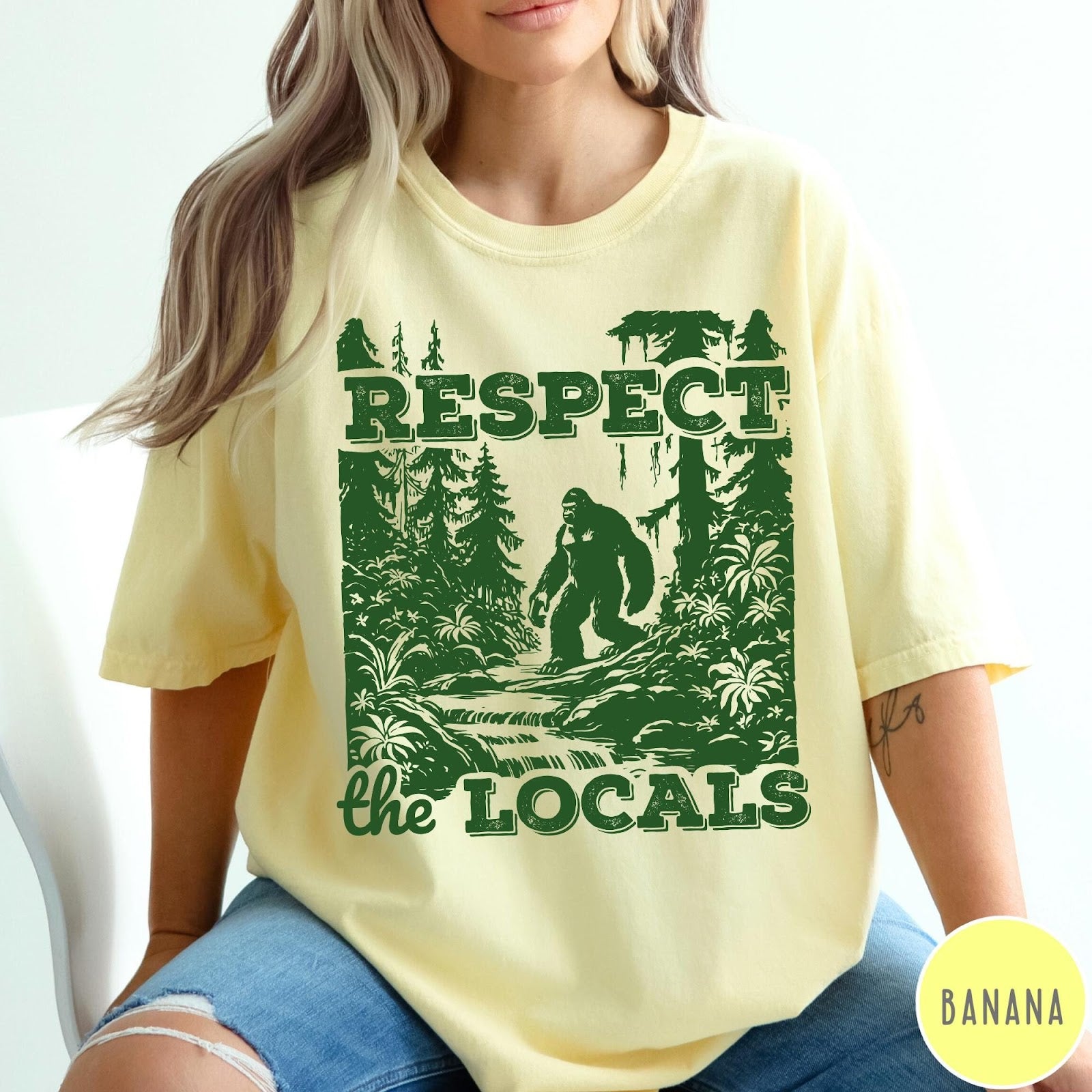 Comfort Colors tee in Banana featuring vintage style graphic of Bigfoot walking through a forest by a river