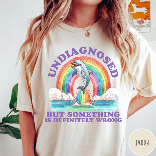 Comfort Colors tee in Ivory featuring graphic style illustration of a dolphin jumping through a colorful rainbow