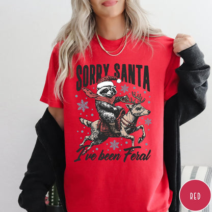 Comfort Colors tee in Red featuring festive graphic of a raccoon wearing a Santa hat riding a reindeer