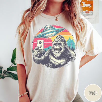 Comfort Colors tee in Ivory featuring retro Bigfoot selfie graphic with a UFO and sunset mountain background