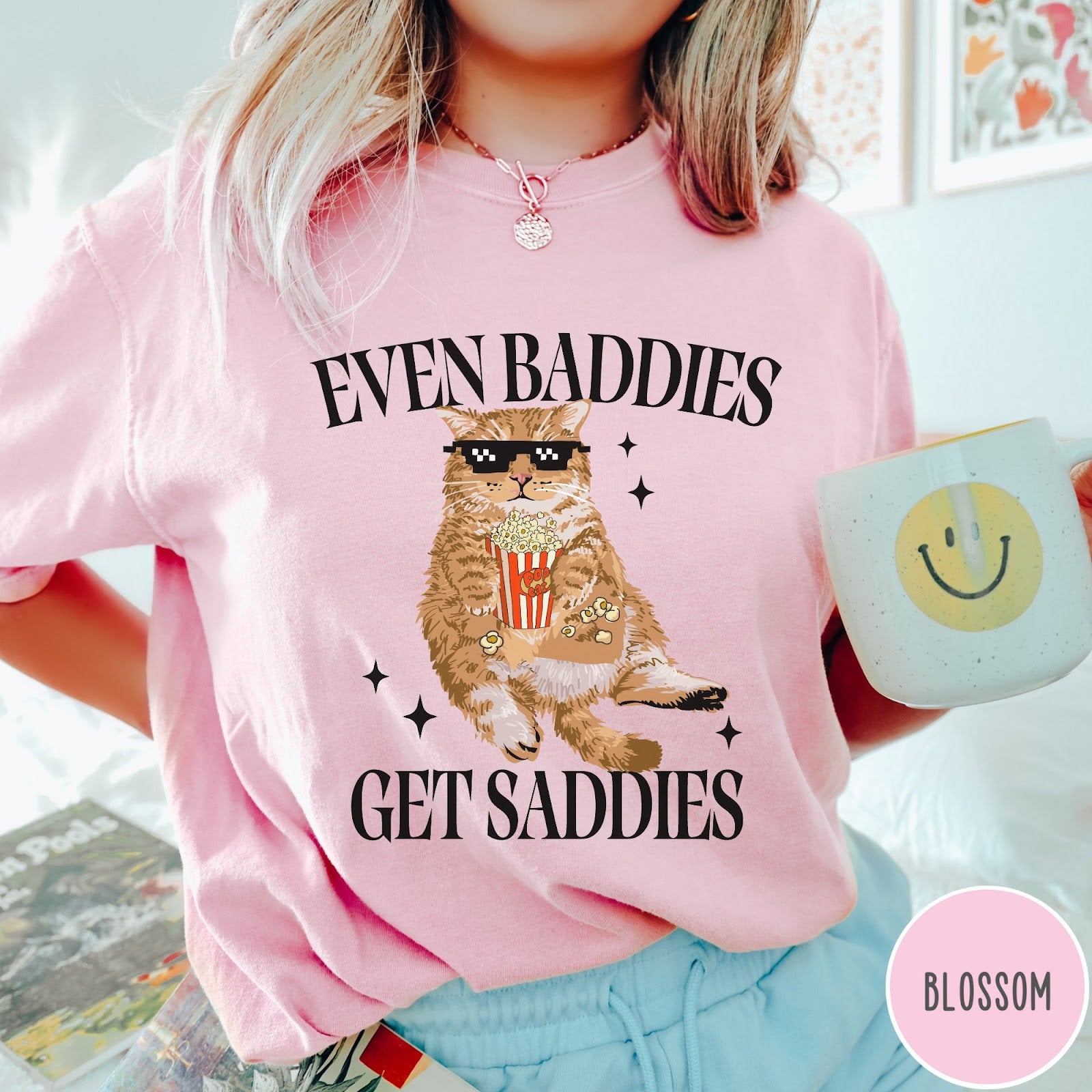 Comfort Colors tee in Blossom featuring cat graphic with sunglasses and popcorn, text Even Baddies Get Saddies