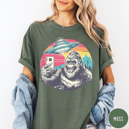 Comfort Colors tee in Moss featuring retro Bigfoot selfie graphic with a UFO and sunset mountain background