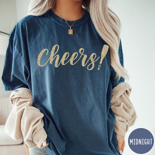 Comfort Colors tee in Midnight featuring New Year celebration graphic with gold glitter cheers text and champagne bottle