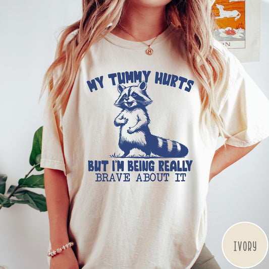Comfort Colors tee in Ivory featuring graphic illustration of a standing raccoon with funny text about being brave