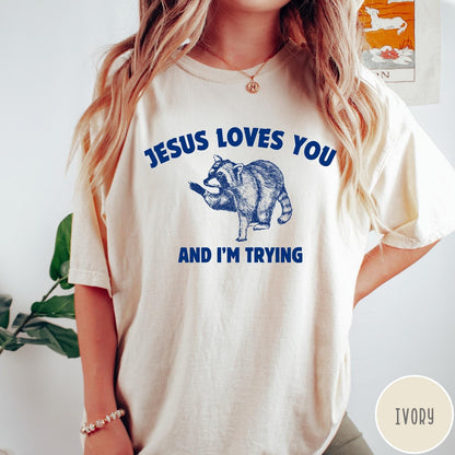 Comfort Colors tee in Ivory featuring graphic style raccoon illustration with text Jesus Loves You And I'm Trying